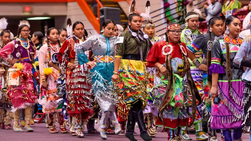 A diverse and vibrant group of enthusiastic individuals, hailing from various backgrounds and cultures, joyfully stride together in perfect unison along the bustling and lively streets, in close proximity to one another, during the mesmerizing and awe-inspiring spectacle of the renowned Black Hills Powwow Event Rapid City