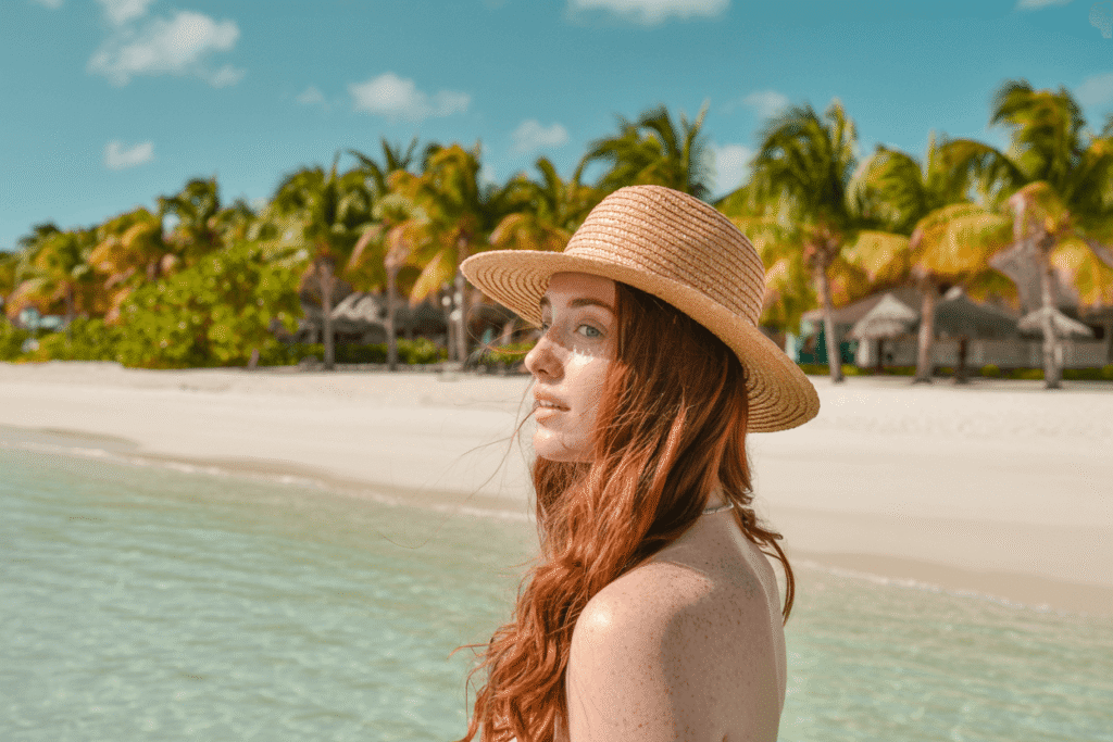A young woman with vibrant red hair and freckled skin stands on a pristine, white-sand beach—one of the best warm October getaways. She is wearing a wide-brimmed straw hat, casting a shadow across her face, and a delicate chain around her neck. Lush palm trees and overwater bungalows dot the background, set against a clear blue sky and turquoise water.